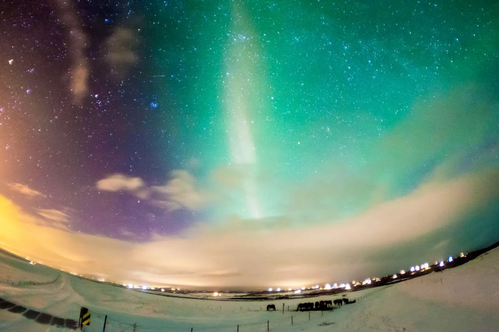 Aurora breaking through between cloud banks over village lights — nights like this are decided by the vedur.is cloud map