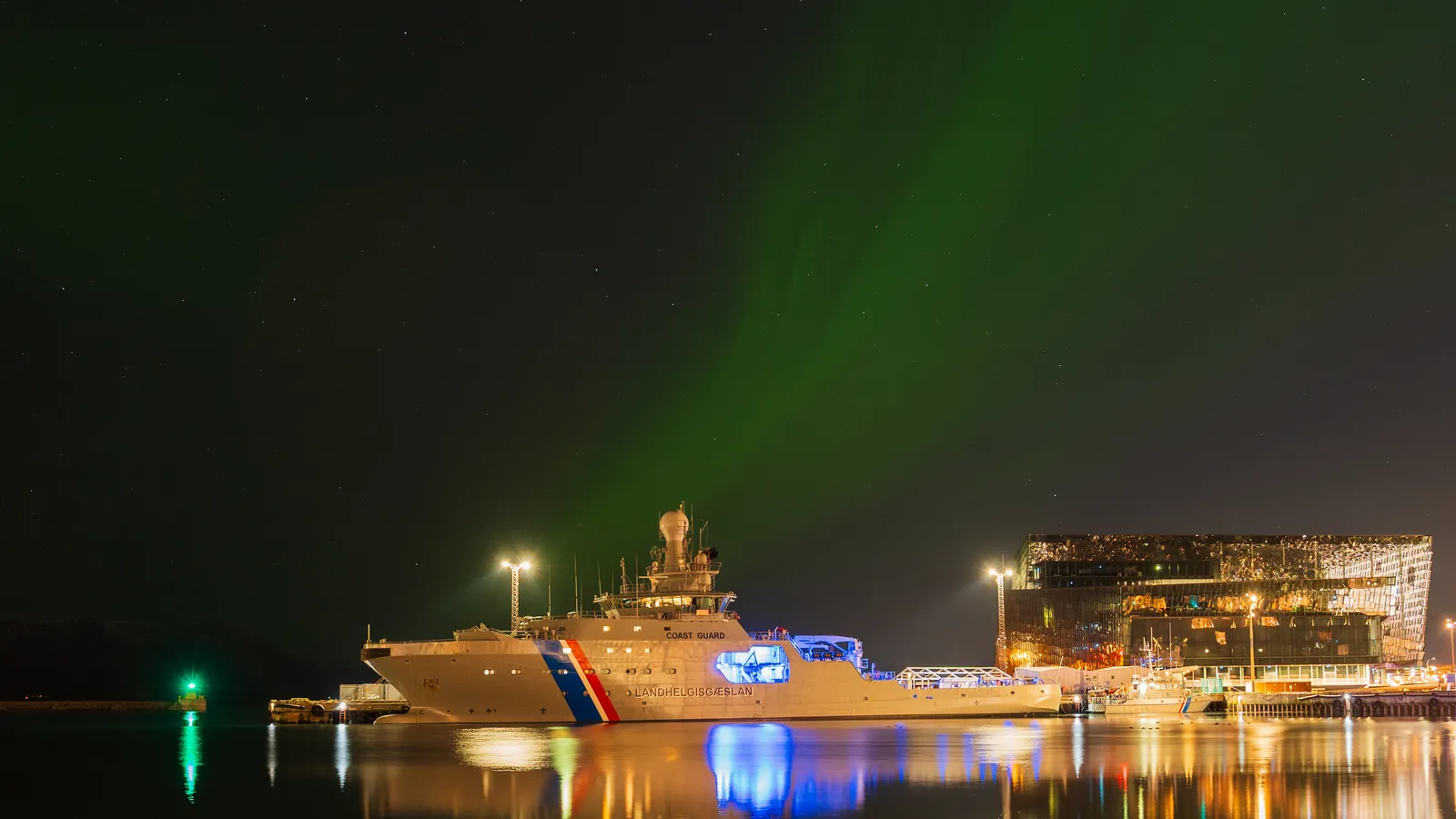 Northern lights over Reykjavík's harbor beside Harpa — on clear nights the aurora is visible from the city itself